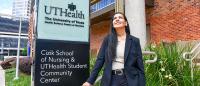 Shabana Momin in business attire stands smiling outdoors beside a sign for UTHealth The University of Texas Health Science Center at Houston Cizik School of Nursing and Student Community Center, with brick walls, and greenery in the background.
