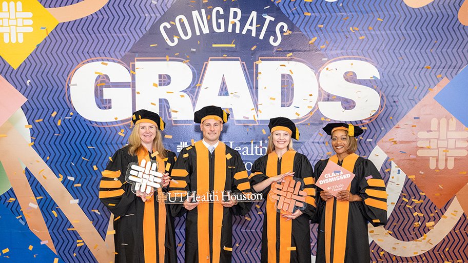 Four graduates in black and orange academic regalia pose together in front of a colorful “Congrats Grads” backdrop at a UTHealth Houston graduation celebration, holding graduation-themed props as confetti covers the floor.
