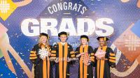 Four graduates in black and orange academic regalia pose together in front of a colorful “Congrats Grads” backdrop at a UTHealth Houston graduation celebration, holding graduation-themed props as confetti covers the floor.