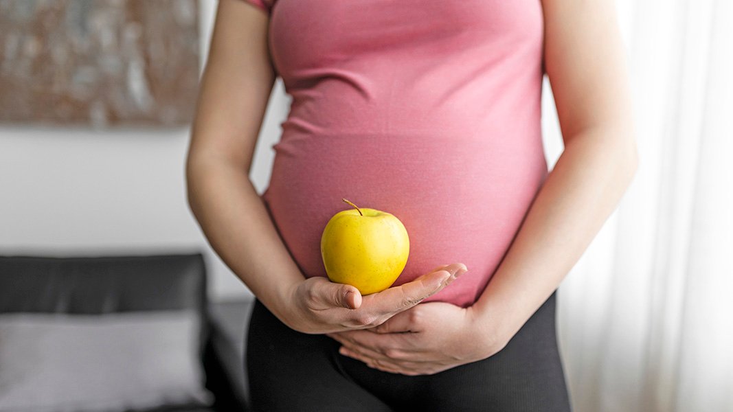 A pregnant person wearing a pink top and black pants holds a yellow apple against their belly with both hands.