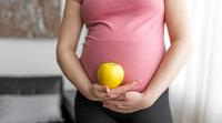 A pregnant person wearing a pink top and black pants holds a yellow apple against their belly with both hands.