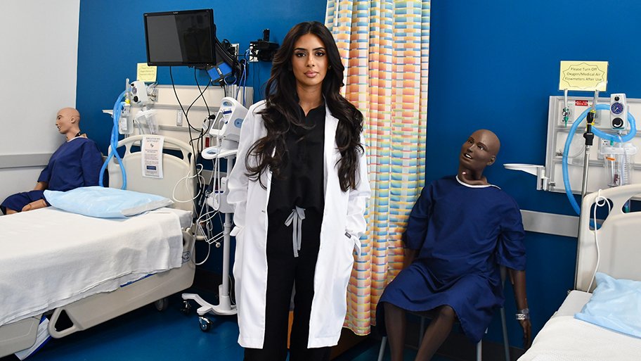FNP student Ali Anjiya stands in the Cizik School of Nursing simulation lab wearing a white coat, surrounded by medical equipment, hospital beds, and patient mannequins.