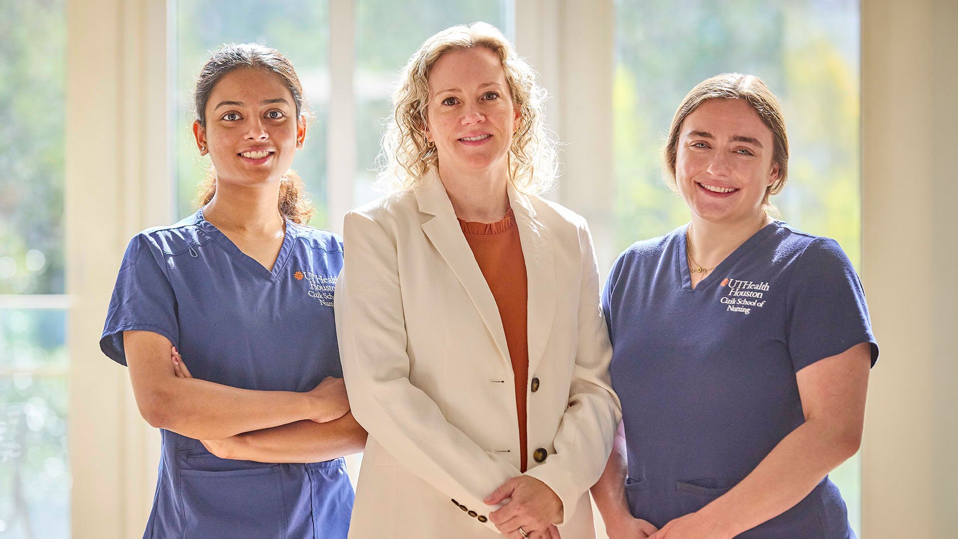 Three people stand side by side indoors in front of large sunlit windows. Two people wear blue UTHealth Houston Cizik School of Nursing scrubs, while the person in the center wears a light-colored blazer and shirt.