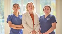 Three people stand side by side indoors in front of large sunlit windows. Two people wear blue UTHealth Houston Cizik School of Nursing scrubs, while the person in the center wears a light-colored blazer and shirt.