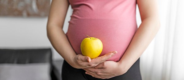 A pregnant person wearing a pink top and black pants holds a yellow apple against their belly with both hands.