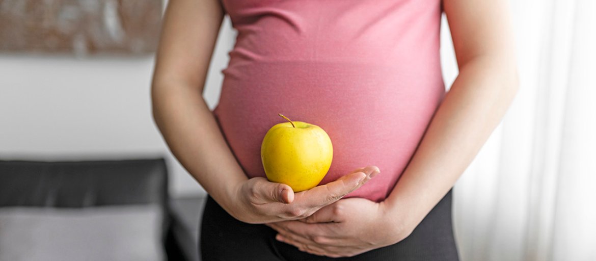 A pregnant person wearing a pink top and black pants holds a yellow apple against their belly with both hands.